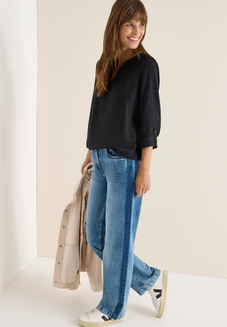 Frau mit schwarzer Bluse, blauer Jeans und Sneakers hält eine helle Jacke.